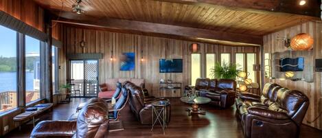 A spacious living area with leather sofas, a coffee table, and a TV. Large windows offer a view of the outdoors.