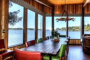 Un coin repas en bois avec une grande table et des chaises, de grandes fenêtres donnant sur un lac et un luminaire suspendu.