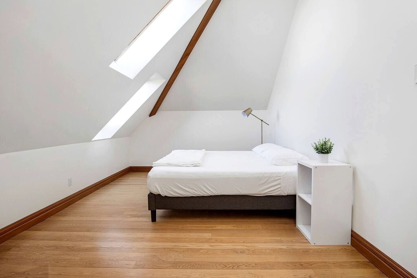 A modern, minimalistic bedroom with a sloped ceiling, a bed, a bedside table with a lamp, and a small plant.