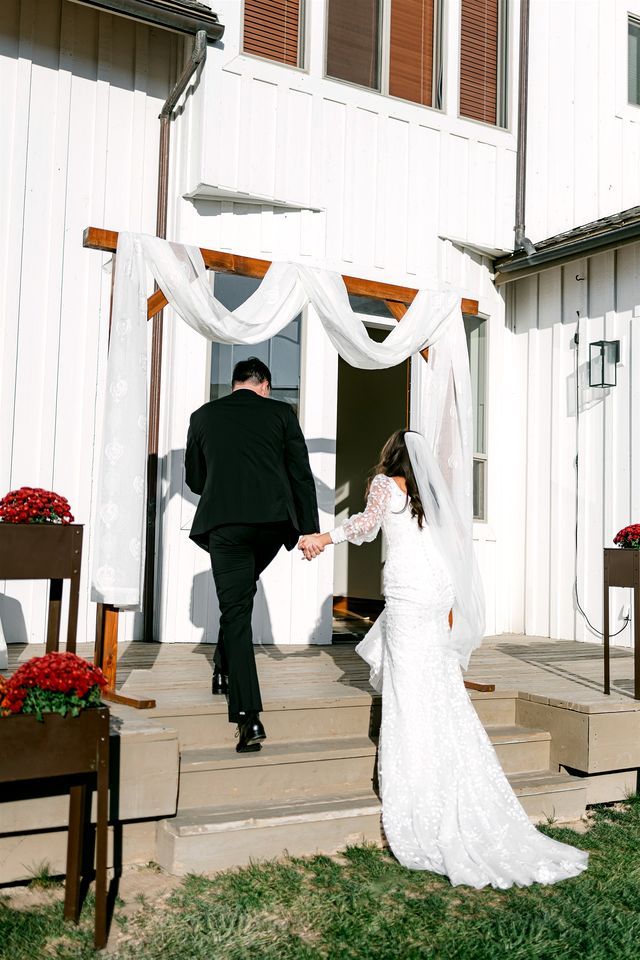 A couple walking towards a white building with a wooden bench and red flowers nearby.