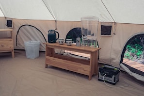 A tent interior with a wooden table, a water dispenser, cups, a kettle, a trash bin, and a power strip.