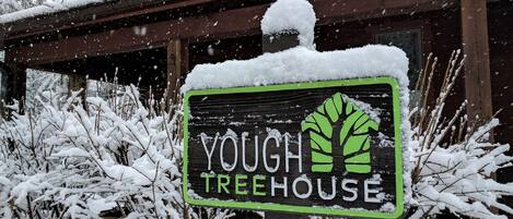 A sign for 'Yough Treehouse' with a green logo, set against a snowy backdrop with a wooden building.