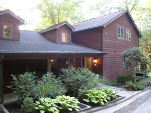 A two-story wooden house with a covered porch, a small attached garage, and a well-maintained garden with various plants.