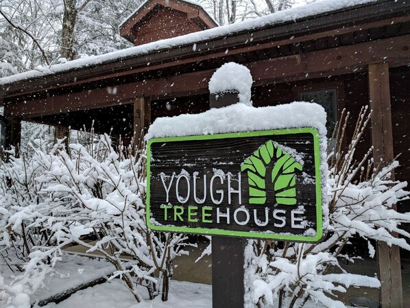 A sign for 'Yough Treehouse' with a green logo, set against a snowy backdrop with a wooden building.