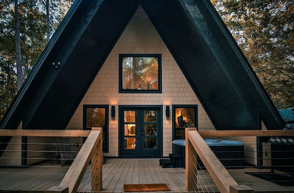 Une cabane triangulaire en bois, dotée d’une fenêtre centrale, d’une porte d’entrée et d’une terrasse avec un banc et un barbecue.