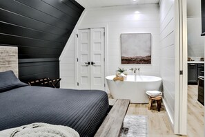 A bedroom with a bed, a small stool, a white bathtub, and a painting on the wall.