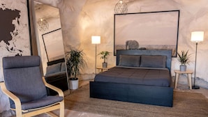 A bedroom with a bed, a chair, a bedside table, a lamp, and a potted plant.