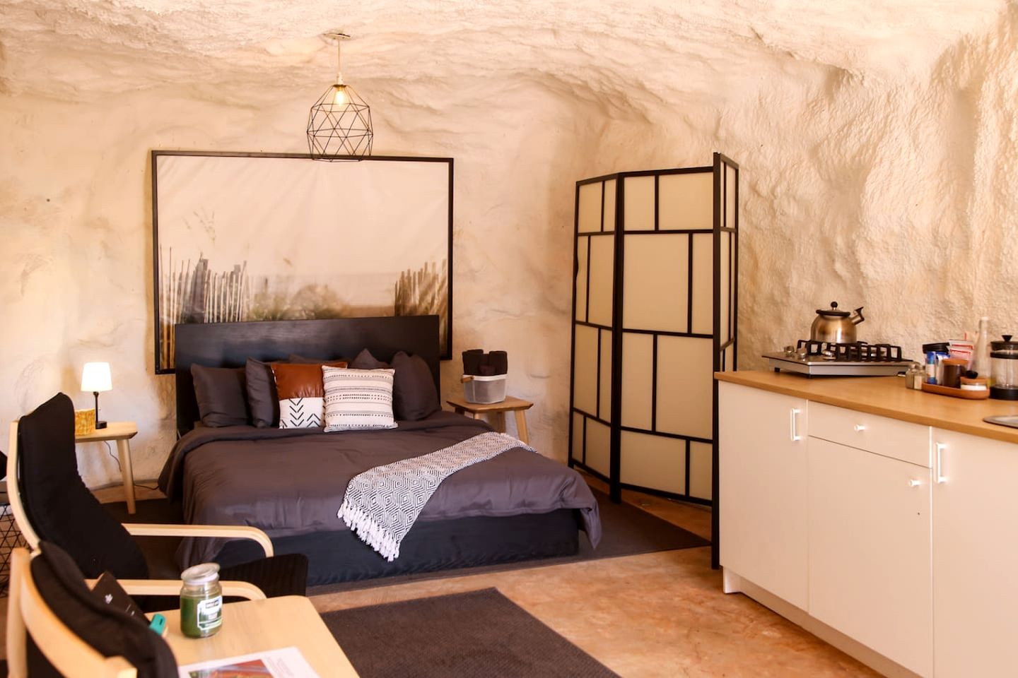 A cozy bedroom with a bed, bedside table, lamp, and a view of a kitchen area.