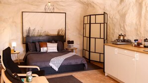 A cozy bedroom with a bed, bedside table, lamp, and a view of a kitchen area.
