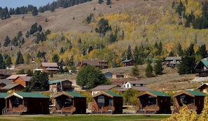 A cluster of wooden cabins with green roofs, set against a backdrop of a hilly landscape with trees and a clear sky. - Beautiful Lakefront Cabin with Stunning Sights in Montana (West Yellowstone)