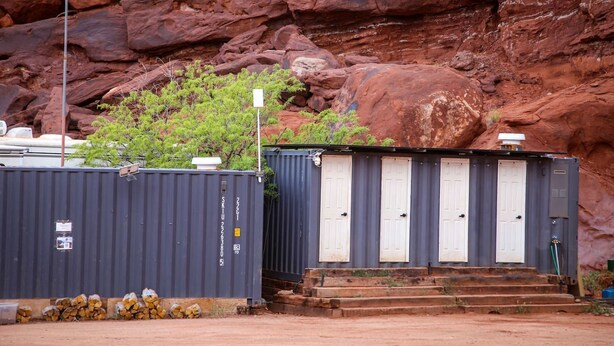 A portable restroom with a white door and a grey exterior, situated in a rocky outdoor setting with a large rock formation in the background. - Incredible Cave for an Unforgettable Glamping Getaway near Canyonlands National Park in Moab, Utah (Moab)