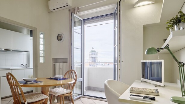 A modern kitchen with white cabinets, a dining table with chairs, a large window with a view of a building, and a desk with a computer and lamp.