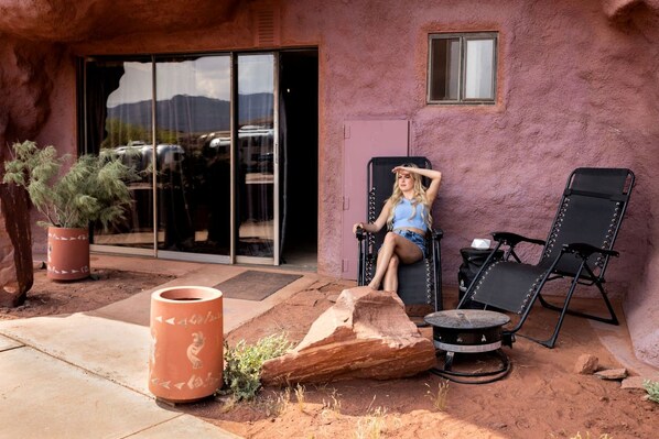 A person sitting on a lounge chair outside a building with a large rock, a potted plant, and a small window.