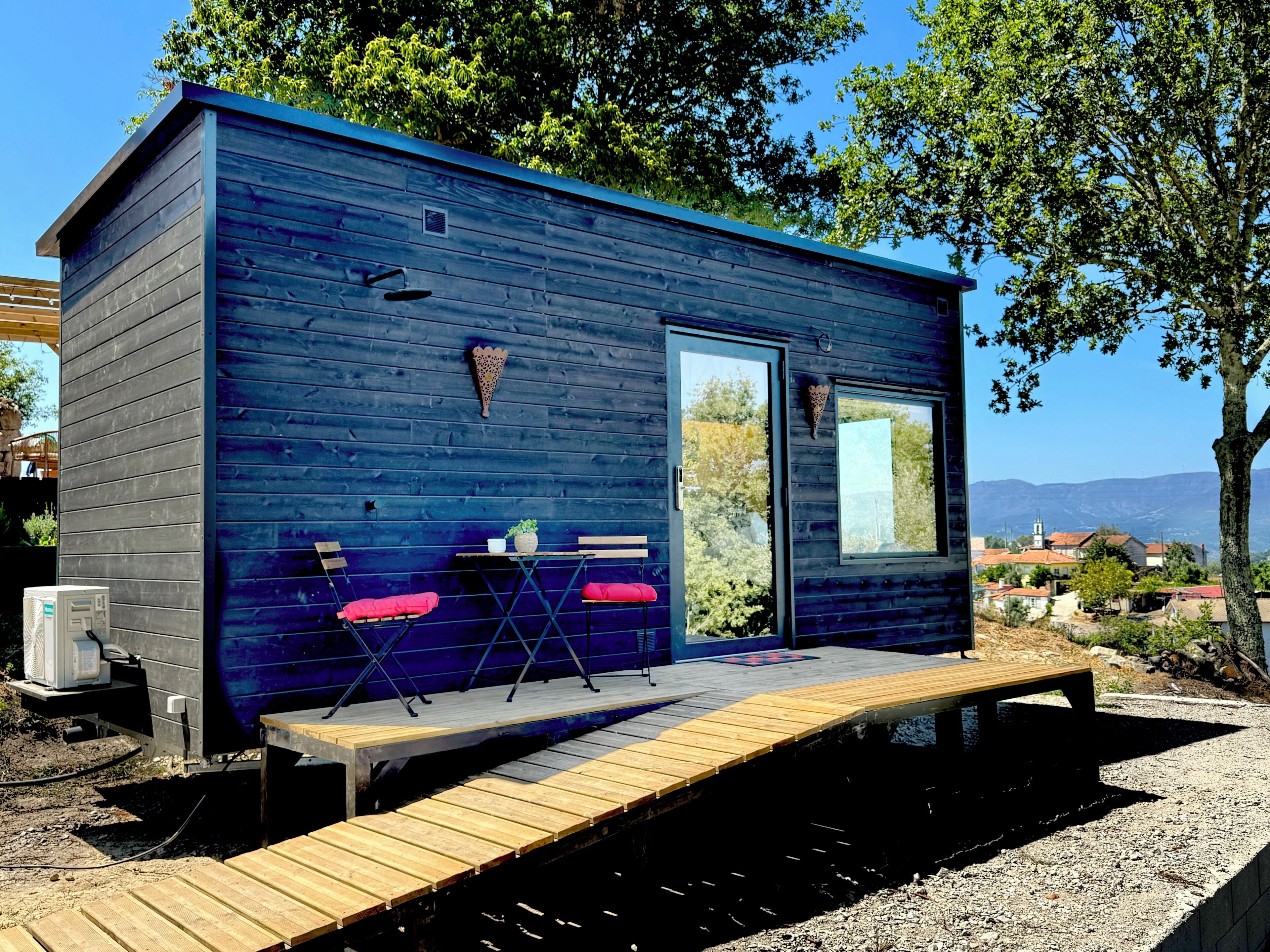 Una pequeña cabaña de madera con terraza, dos sillas, una mesa y un ventanal.