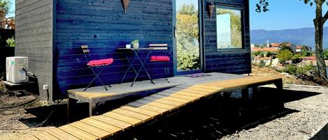 Una pequeña cabaña de madera con terraza, dos sillas, una mesa y un ventanal.