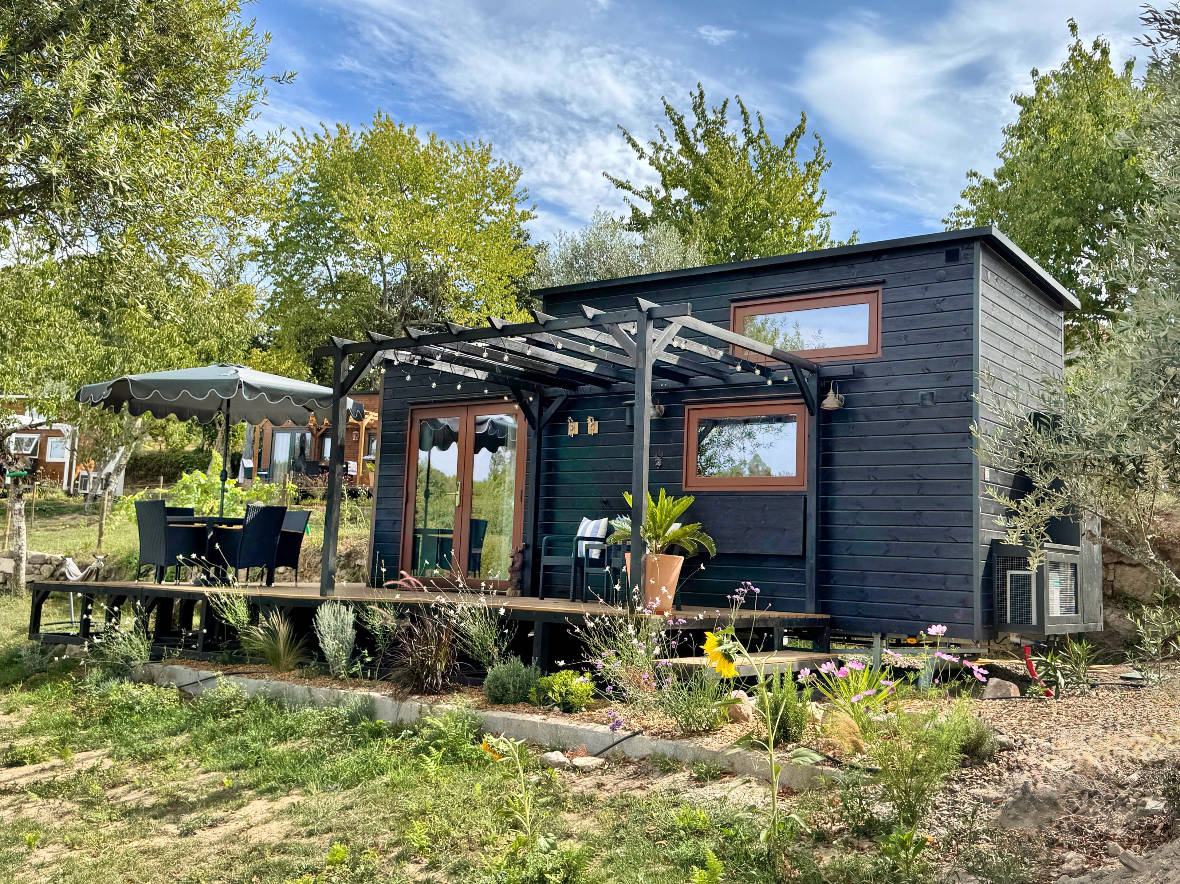 A small, dark-colored cabin with a covered patio area, outdoor seating, and a garden with various plants and flowers.