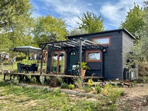 A small, dark-colored cabin with a covered patio area, outdoor seating, and a garden with various plants and flowers. - Tiny Escape (Vouzela)