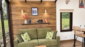 Ein grünes Sofa mit gemusterten Kissen, ein Holztisch, eine Wandablage mit Dekoration und ein Fenster mit Blick auf Grünflächen.