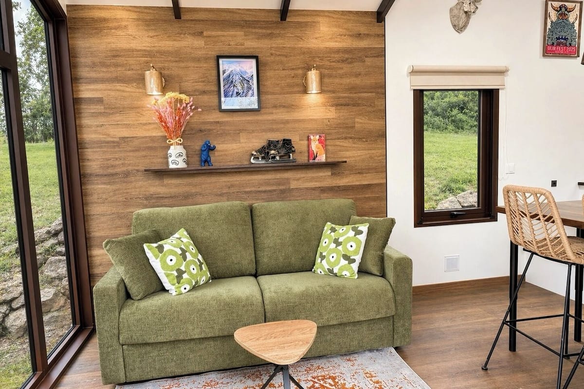 Ein grünes Sofa mit gemusterten Kissen, ein Holztisch, eine Wandablage mit Dekoration und ein Fenster mit Blick auf Grünflächen.