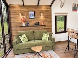 Un sofá verde con almohadones estampados, una mesa de centro de madera, un estante en la pared con adornos, y una ventana con vista a la vegetación.