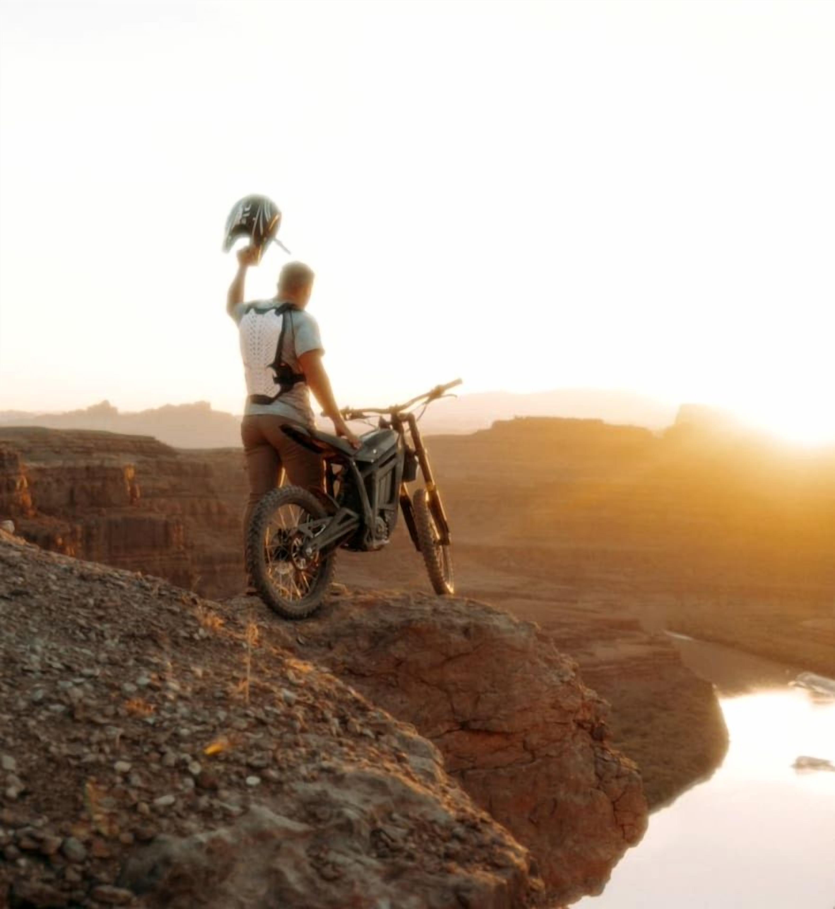 Eine Person auf einem Motorrad fährt über steiniges Gelände, im Hintergrund ein Sonnenuntergang.