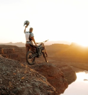 Una persona en motocicleta sobre terreno rocoso, con un atardecer de fondo.