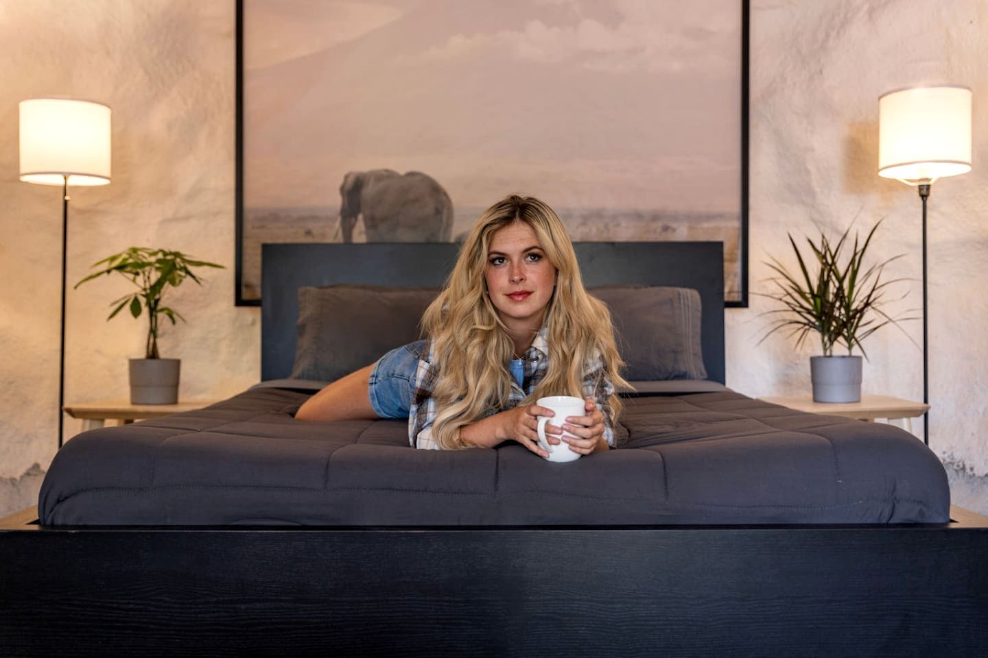 A bed with a dark mattress, a person holding a mug, two bedside lamps, and a framed picture of an elephant on the wall.