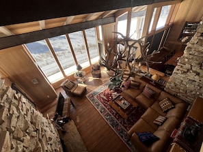 A spacious living room with a large window, a stone wall, and a wooden ceiling.