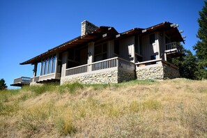 A two-story stone house with a wooden porch and large windows, situated on a grassy hill.