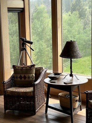 A room with a large window, a wicker chair, a table with a lamp, and a telescope on a tripod.