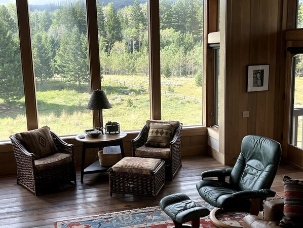 A cozy room with large windows, wicker furniture, a leather chair, and a small table with a lamp.