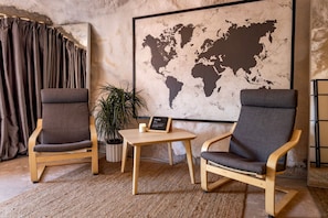 Een kamer met twee houten stoelen, een klein tafeltje, een potplant, een wereldkaart aan de muur en een gordijn.