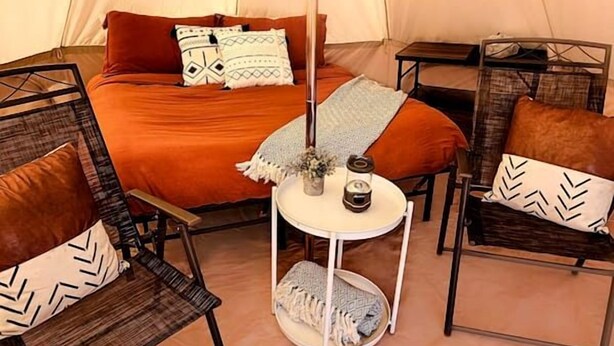 Een tent met een bed, twee stoelen, een kleine tafel met een kaars, en een vaas met bloemen.