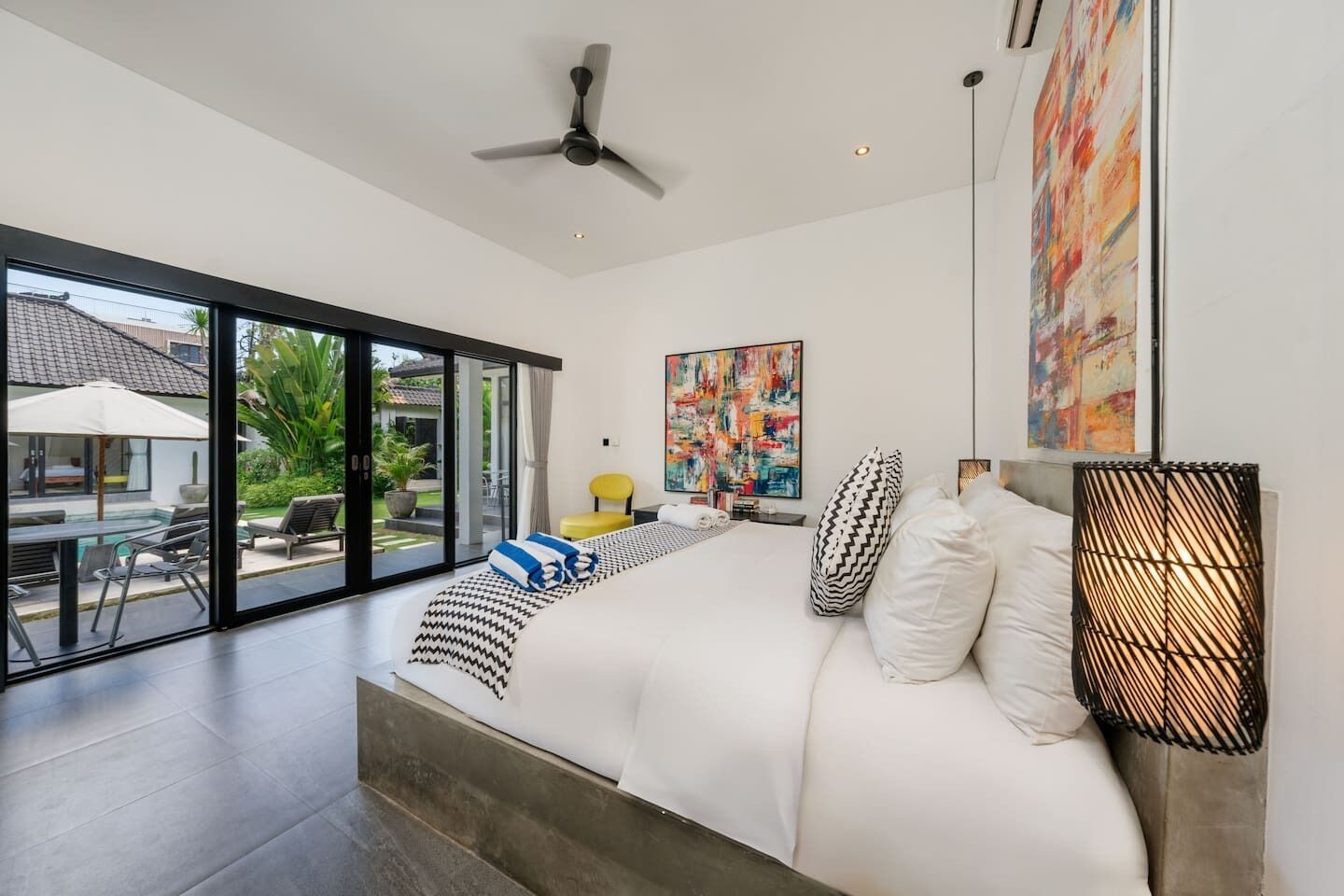 A modern bedroom with a large bed, a ceiling fan, and a view of a swimming pool and outdoor area.