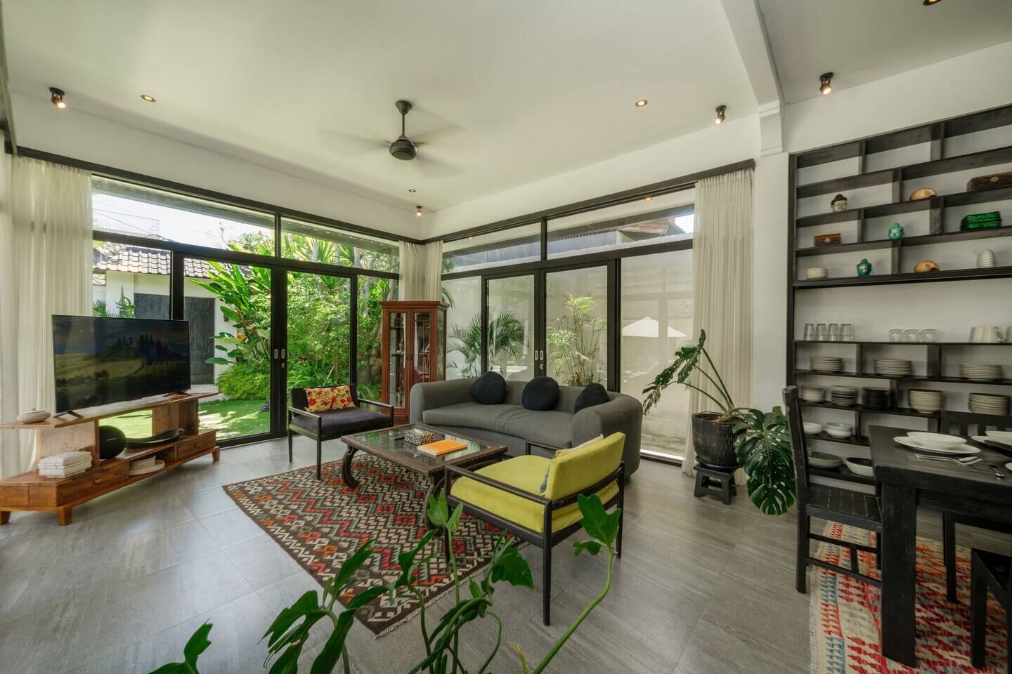 A spacious living room with a large TV, comfortable seating, and a dining area with a shelf of dishes.