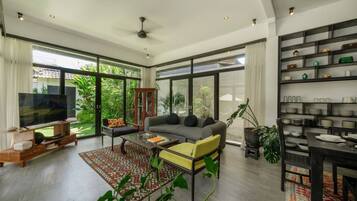 A spacious living room with a large TV, comfortable seating, and a dining area with a shelf of dishes.