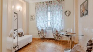 A room with a white sofa, a glass table, a chair, a floral curtain, a clock, and a framed picture on the wall.