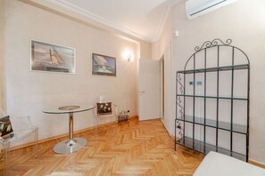 Une pièce dotée d’un parquet en bois à motifs chevrons, d’une table en verre avec un pied en métal, de deux tableaux encadrés accrochés au mur, d’une porte en métal et d’une chaise en plastique transparent.