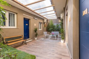 Un espacio al aire libre cubierto con un banco, sillas, una mesa, plantas en macetas y una puerta azul.