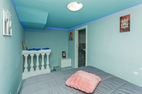 Un dormitorio con una cama, una mesita de noche blanca, una puerta e iluminación LED azul en el techo.