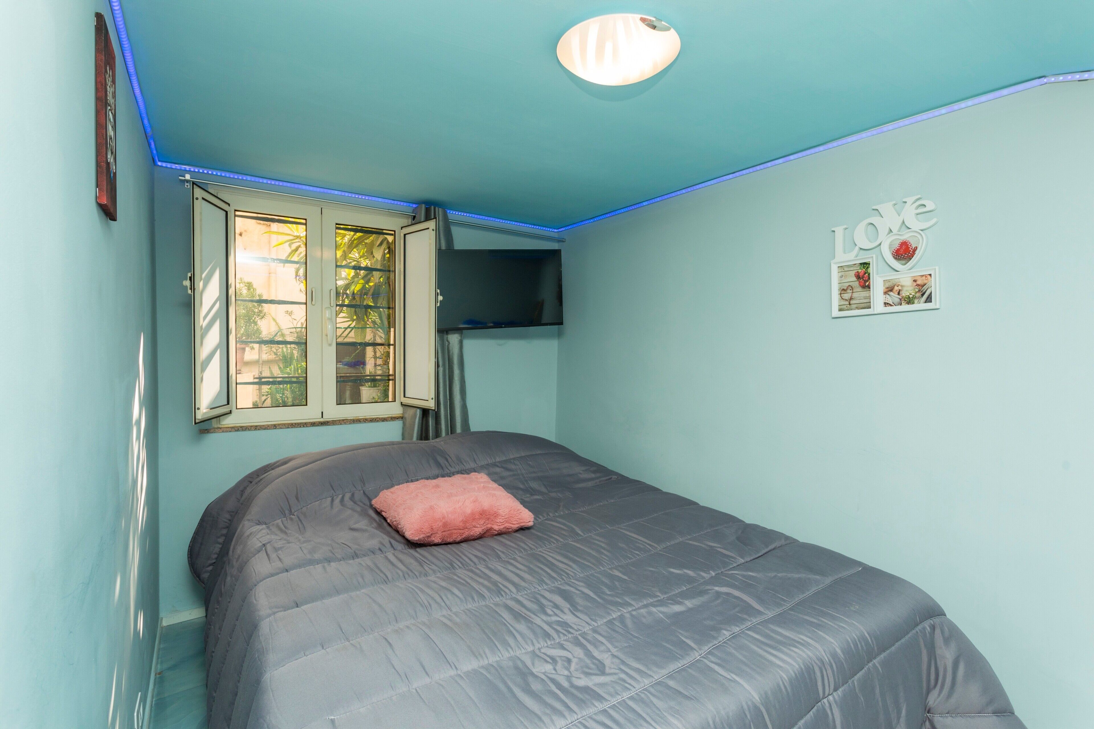 Een slaapkamer met een bed, een raam, een aan de muur bevestigde televisie en een plafondlamp.