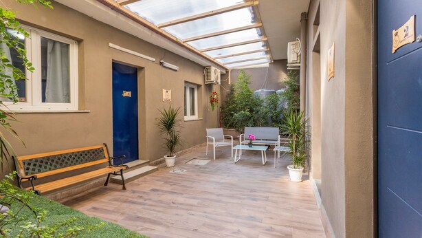 Un espacio al aire libre cubierto con un banco, sillas, una mesa, plantas en macetas y una puerta azul.