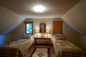 A small, cozy attic bedroom with two single beds, a table with lamps, a mirror, and a window with blinds. - Charming Riverside Cabin with Deck Overlooking Madison River near Three Dollar Bridge in McAllister, Montana (Cameron)