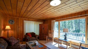 Interior acogedor de una cabaña con techo de madera, un ventanal con vistas a árboles, chimenea y un espacio de descanso cómodo con un sofá y sillones.