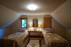 A small, cozy attic bedroom with two single beds, a table with lamps, a mirror, and a window with blinds.