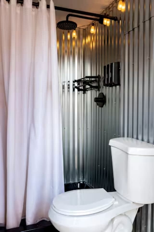 A bathroom with a white toilet, a shower with a black hose, and a metal wall.