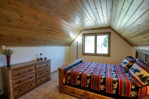 A bedroom with a sloped ceiling, a wooden dresser, a bed with patterned bedding, and a window with a view of trees.