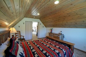 A bedroom with a sloped ceiling, a bed with a patterned blanket, a wooden dresser, a chair, and a lamp.