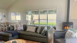 A living room with a sectional sofa, a small round table, a fireplace, and large windows overlooking a backyard. - The Salty Olive - WiFi, Pet Friendly Outside (Robe)