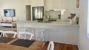 A modern kitchen with white cabinetry, a stainless steel refrigerator, a built-in oven, and a small island with a sink. There are white chairs and a wooden table with placemats. - The Salty Olive - WiFi, Pet Friendly Outside (Robe)